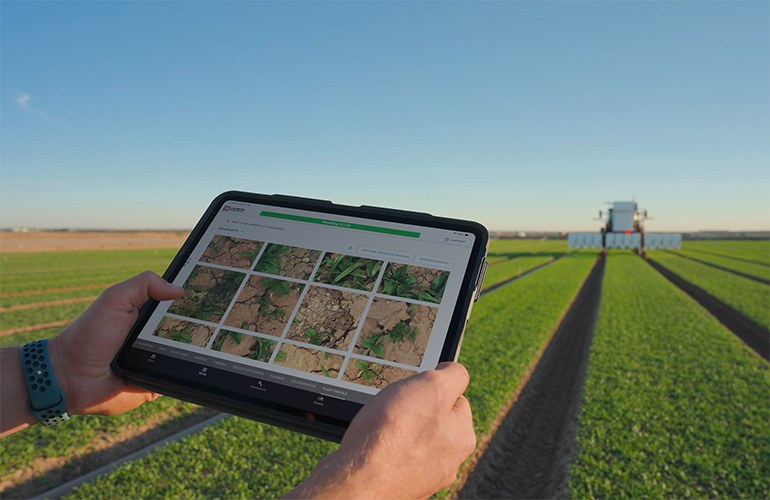 From Lab to Field: Carbon Robotics and iMerit Unleash Instant AI Customization close up of an ipad in foreground with a tractor in a field in the background.