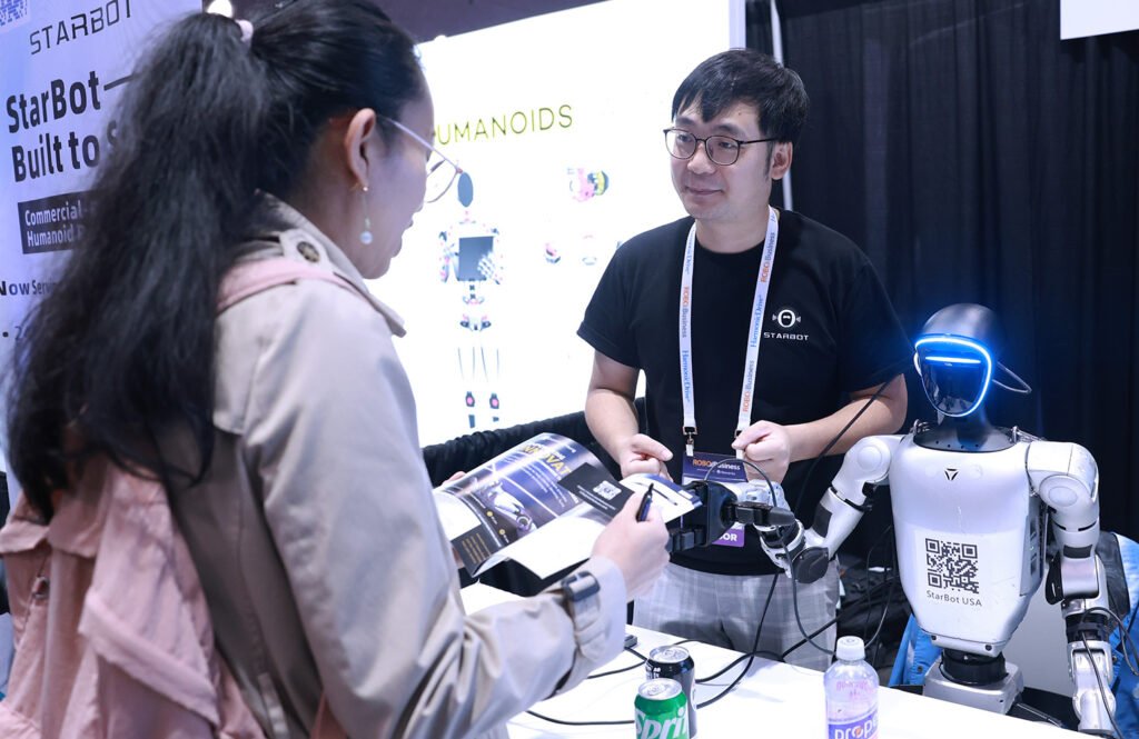 RoboBusiness 2026 Day 1: Everything You Need to Know A woman talking to a man and a humanoid robot at RoboBusiness 2025. RoboBusiness 2026 will feature sessions on humanoids.